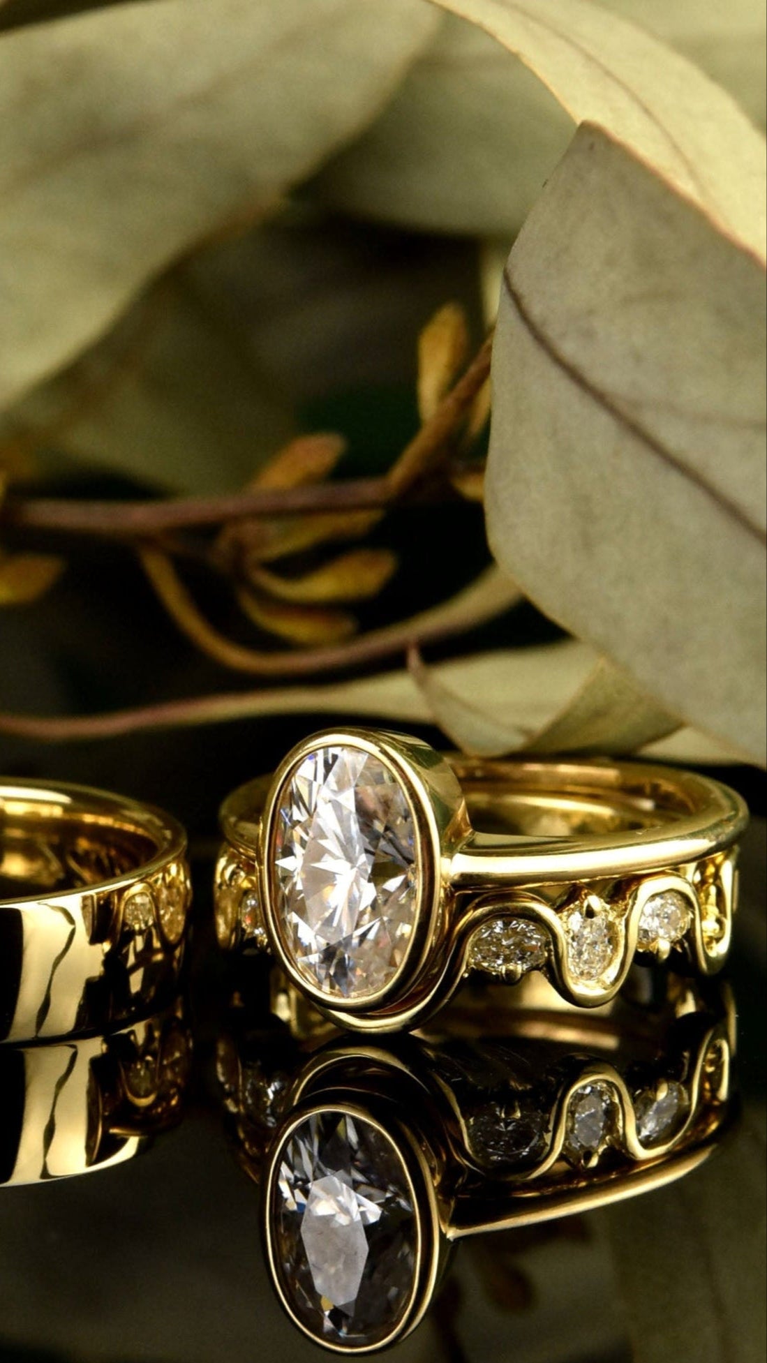 Two gold rings with a diamond on a reflective surface with leaves in the background